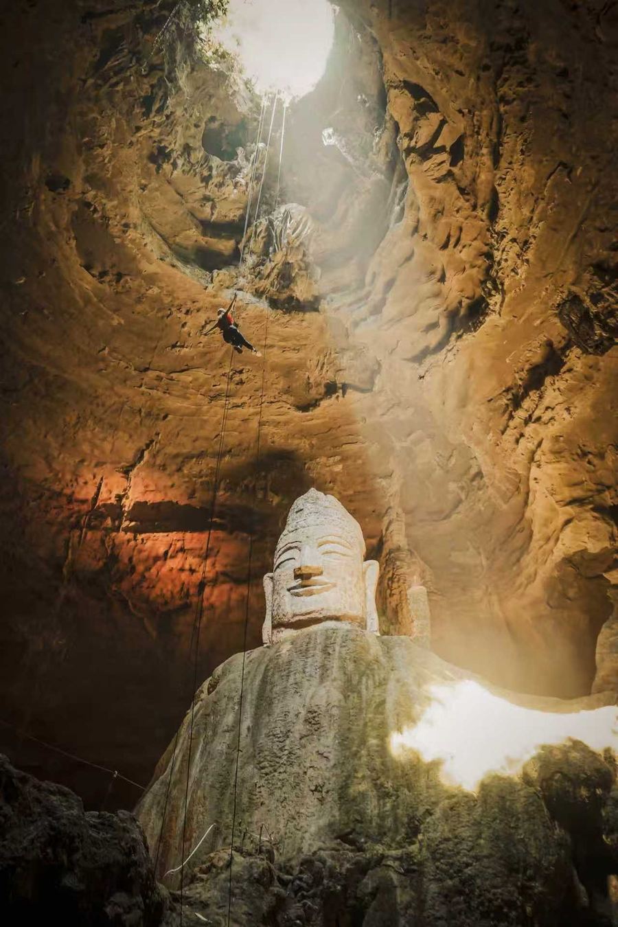 Linda abseiling down a rope through the opening of Tianyan Cave towards a giant stone Buddha